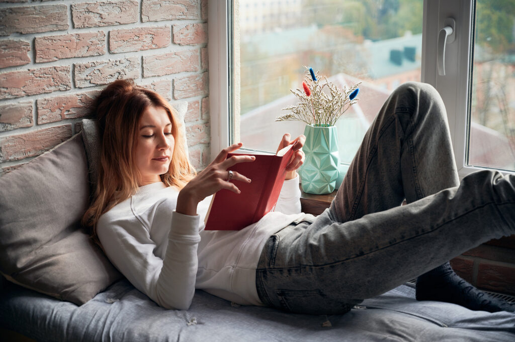 Young attractive woman in white sweatshirt lying and reading interesting book on bed near view from window